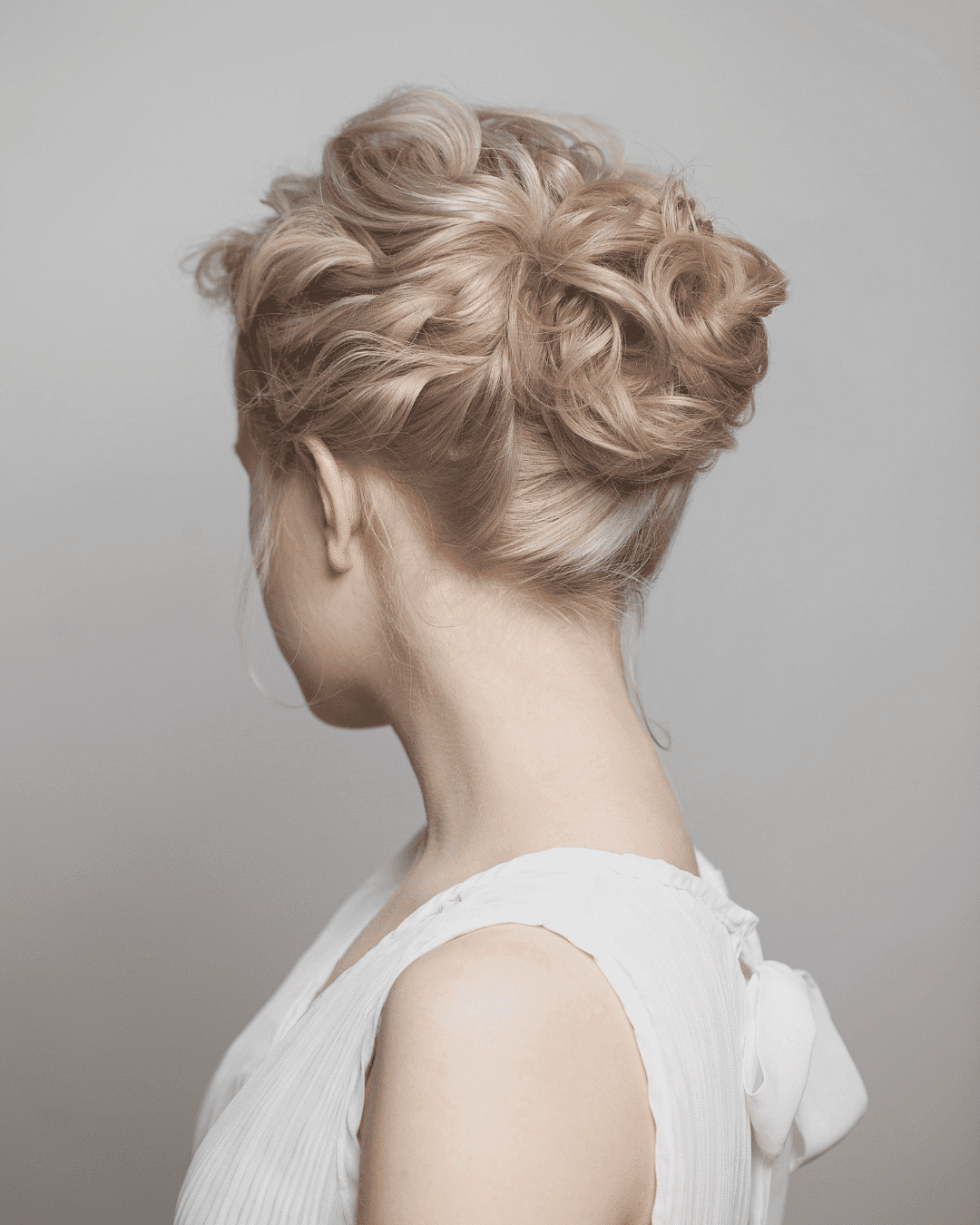 Woman with elegant updo hairstyle, viewed from the side, wearing a white sleeveless top.